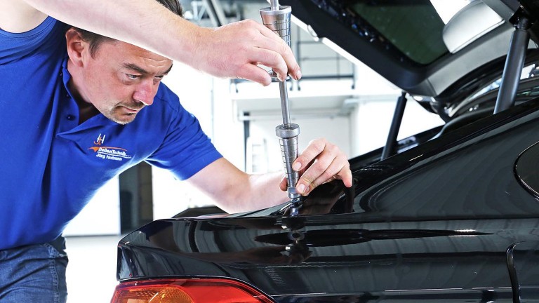 Jörg Hofmann repariert eine Delle im Kofferraumdeckel eines schwarzen Fahrzeugs.