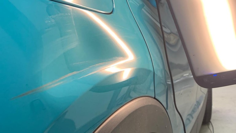 Langer Kratzer im türkisfarbenen Lack des hinteren rechten Kotflügels, sichtbar durch Spiegellicht.